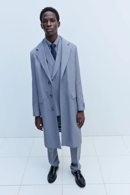 A tall, slender Black man wearing a grey three-piece suit and matching overcoat stands indoors against a light backdrop, fashion shoot.