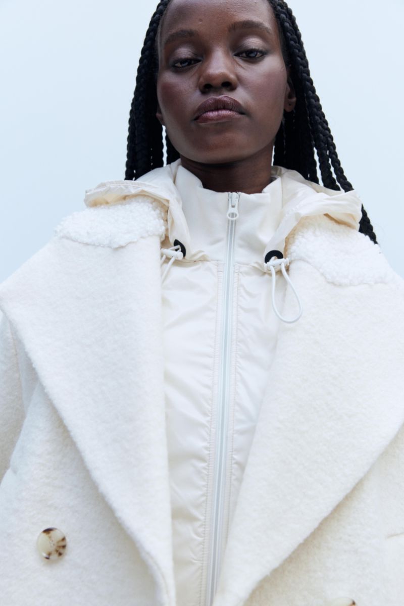 A fashionable woman with braids poses in a photo studio, wearing a cream-colored double-breasted coat over a matching zip-up hooded jacket.