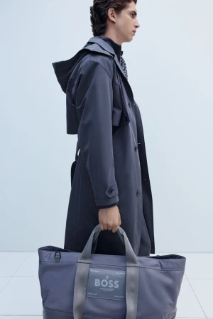 Profile view of a man wearing a dark hooded trench coat and carrying a large grey BOSS tote bag against a plain white background.