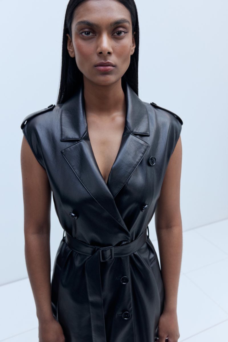 Woman in a black sleeveless, belted leather dress poses in a minimalist studio setting with bright, even lighting. Fashion editorial style.
