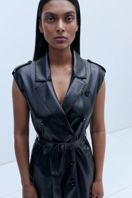 Woman in a black sleeveless, belted leather dress poses in a minimalist studio setting with bright, even lighting. Fashion editorial style.