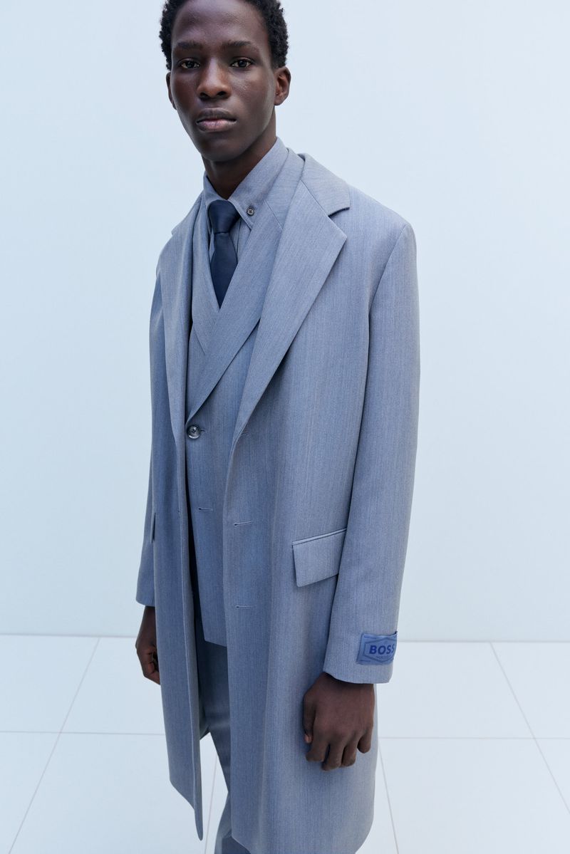 Man in a grey three-piece suit and matching coat, standing indoors against a light wall, showcasing formal fashion.