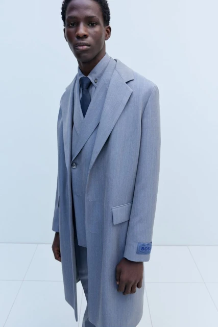 Man in a grey three-piece suit and matching coat, standing indoors against a light wall, showcasing formal fashion.