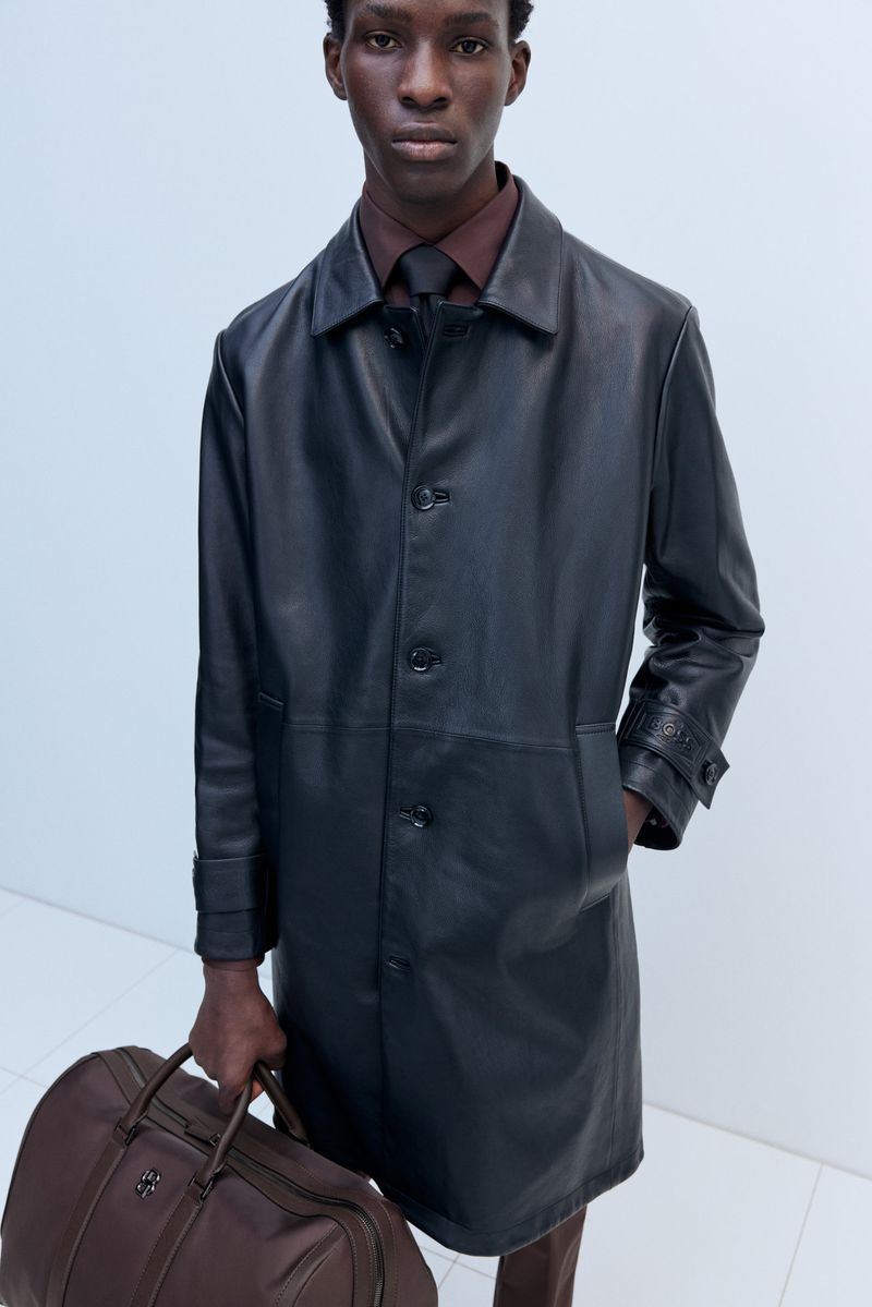 Dark-skinned male model in a black leather trench coat holding a brown leather duffel bag in a minimalist, bright white studio.