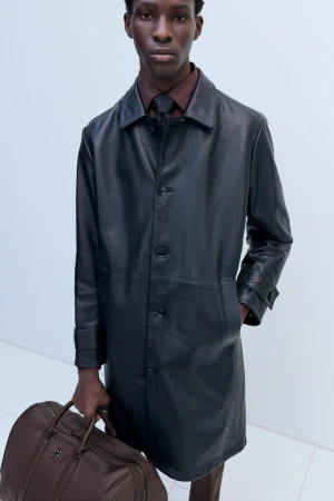 Dark-skinned male model in a black leather trench coat holding a brown leather duffel bag in a minimalist, bright white studio.