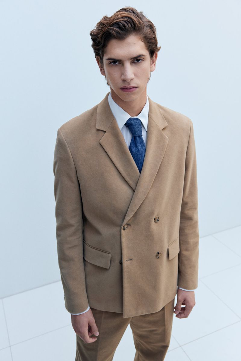 A stylish adult male wearing a tan double-breasted suit with a blue tie stands in front of a plain white background, shot from a high angle.