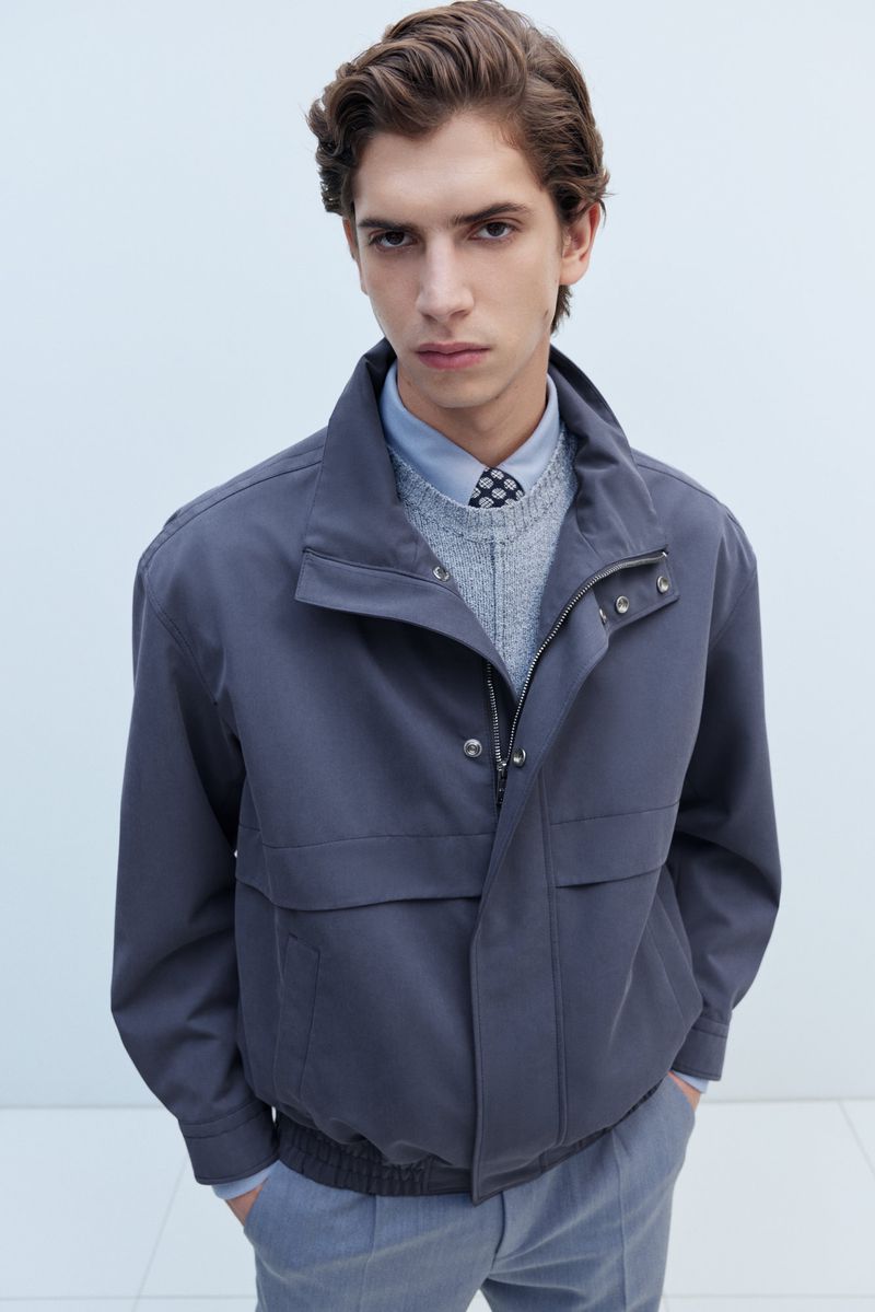 Portrait of a male model wearing a blue-grey jacket over a sweater and shirt in a brightly lit indoor photo studio setting.