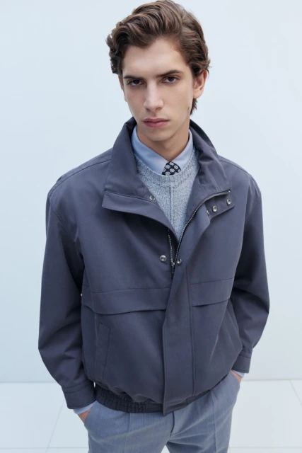 Portrait of a male model wearing a blue-grey jacket over a sweater and shirt in a brightly lit indoor photo studio setting.