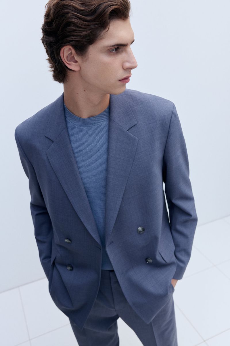 Man in a blue suit and t-shirt looking away from camera in a bright indoor setting with a neutral background in a medium shot.
