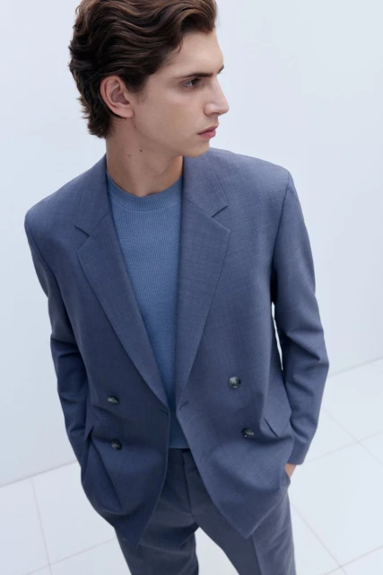 Man in a blue suit and t-shirt looking away from camera in a bright indoor setting with a neutral background in a medium shot.