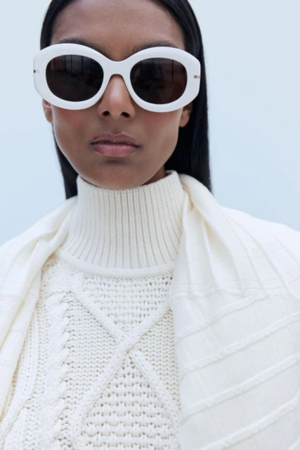A woman poses in white sunglasses, a cable-knit sweater, and a shawl in a close-up studio shot against a light blue background.