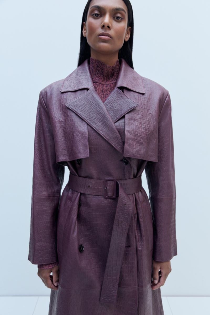 An adult female fashion model poses in a studio wearing a reptile-textured double-breasted trench coat and a brown sweater with a turtleneck.