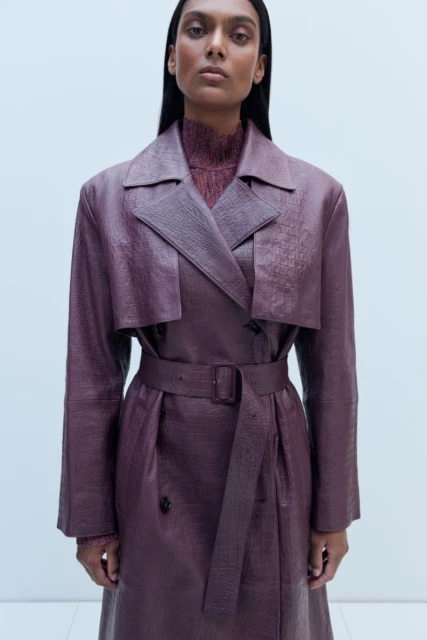 An adult female fashion model poses in a studio wearing a reptile-textured double-breasted trench coat and a brown sweater with a turtleneck.