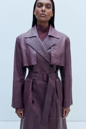 An adult female fashion model poses in a studio wearing a reptile-textured double-breasted trench coat and a brown sweater with a turtleneck.