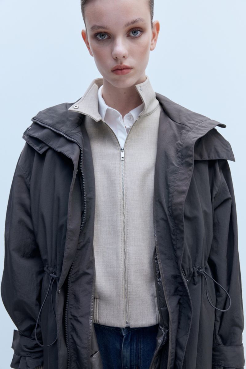 Model wearing a layered dark hooded parka over a beige zip jacket and white collared shirt against a light studio backdrop.