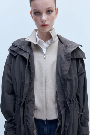 Model wearing a layered dark hooded parka over a beige zip jacket and white collared shirt against a light studio backdrop.