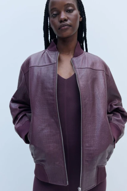 Portrait of an African American woman wearing a burgundy faux-crocodile bomber jacket and dress, posing in a neutral studio setting.