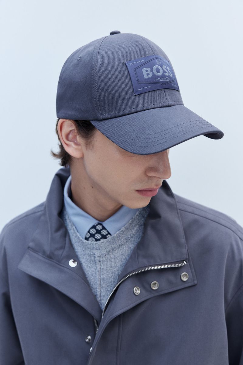 A grey baseball cap with a BOSS logo on the front, worn by a person in a collared shirt, sweater, and jacket, in a studio setting.