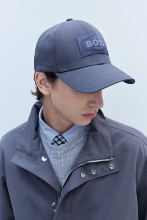 A grey baseball cap with a BOSS logo on the front, worn by a person in a collared shirt, sweater, and jacket, in a studio setting.
