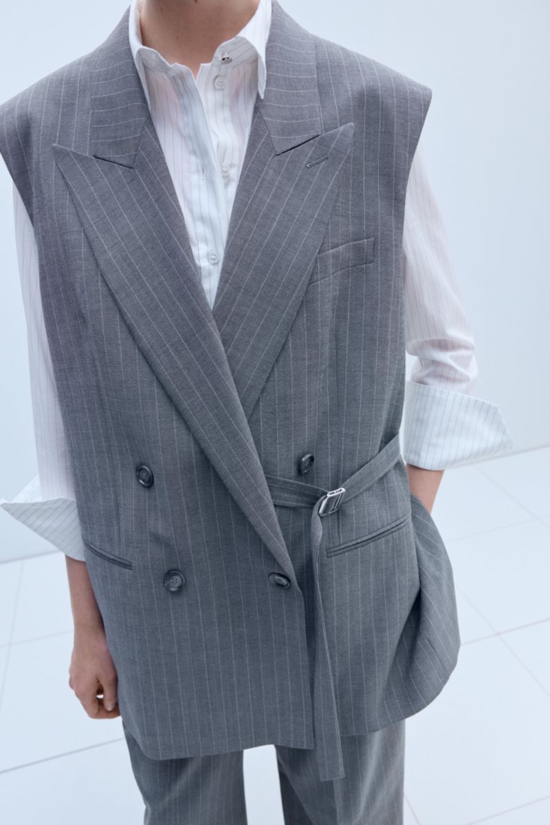 Person wearing a pinstripe sleeveless blazer with a belt, combined with a white striped shirt. Gray suit with double-breasted closure.
