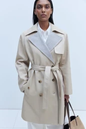Model wearing a beige belted trench coat with a grey lapel and white shirt against a white seamless background