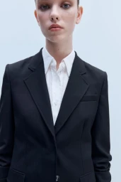 Close-up portrait of a woman wearing a tailored black blazer over a crisp white collared shirt against a pale studio backdrop.