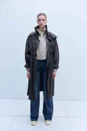 Model standing against a white backdrop wearing a dark coat over a light zip-up jacket and blue jeans.