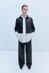Model wearing a black leather jacket and matching pants over a white collared shirt against a white backdrop.