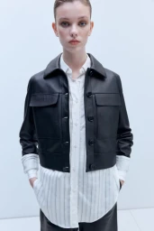Model wearing a cropped black leather jacket layered over a long white pinstripe button-down shirt against a pale studio backdrop.