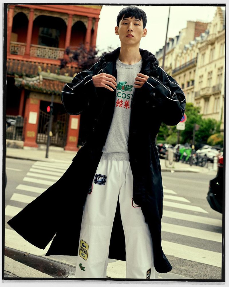 A young, slender man in an open black coat over a Lacoste t-shirt poses on a crosswalk with buildings in the background.