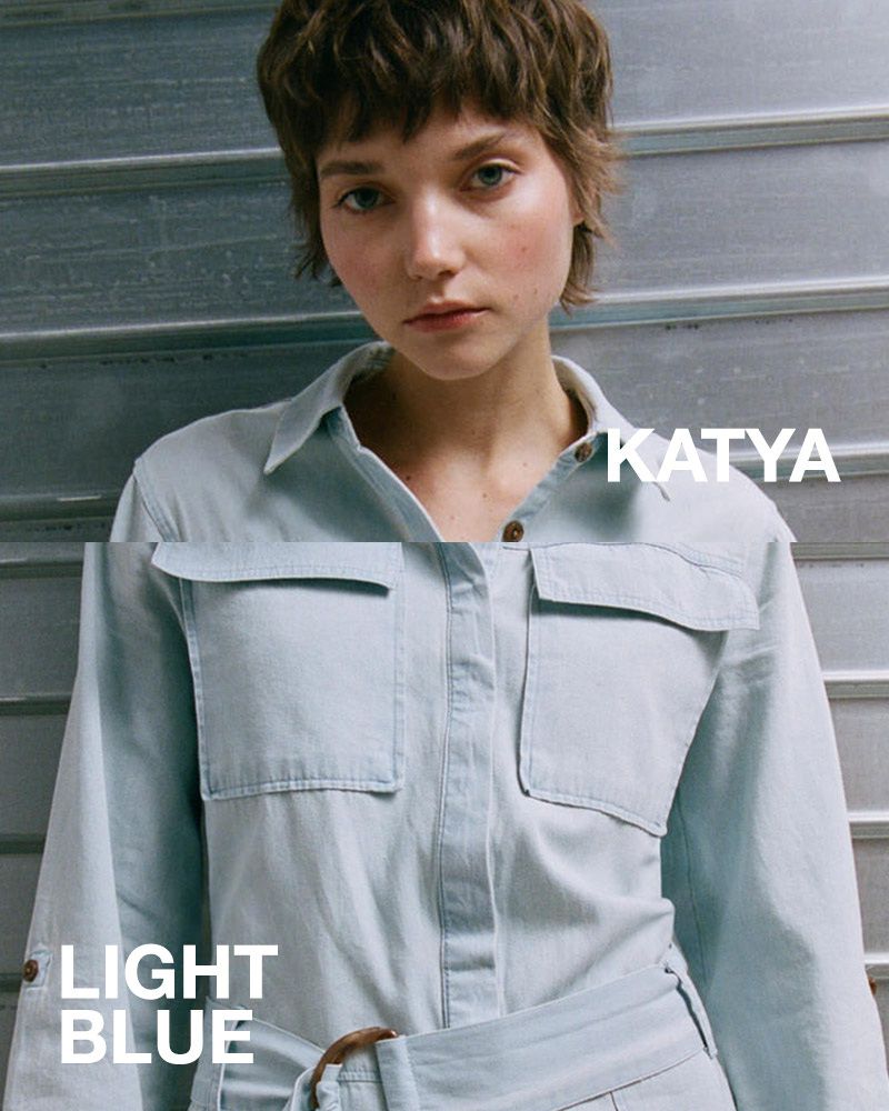 Close-up of a young woman with short brown hair wearing a light blue denim jumpsuit, posing in front of a corrugated metal wall.