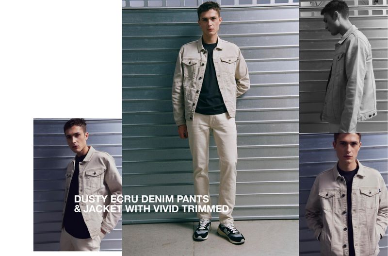 A stylish young man posing in a light beige denim jacket and pants ensemble in front of a corrugated metal backdrop in a fashion editorial.