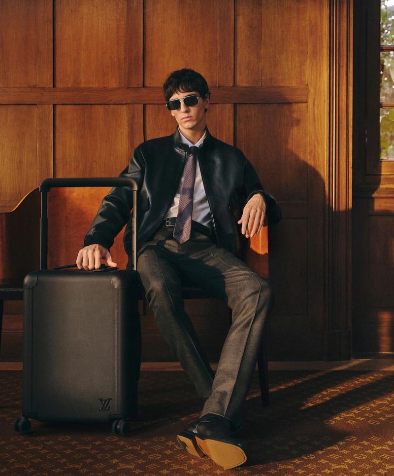 Stylish man sits on a bench next to his black rolling luggage, dressed in a black leather jacket, grey trousers, and sunglasses.