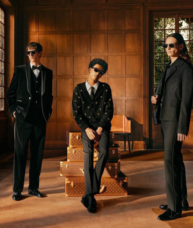 Three young men pose indoors, two in dark suits, one in a cardigan, with vintage Louis Vuitton luggage stacked in the background.