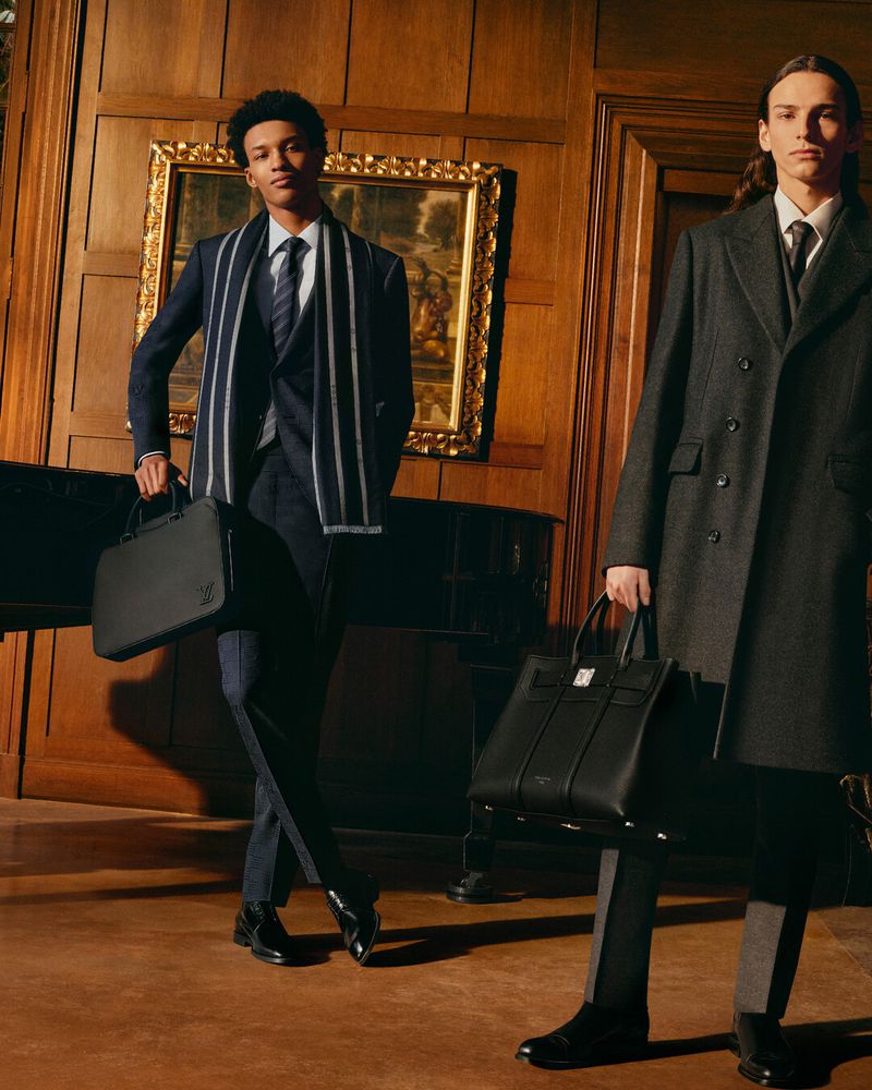 Two adult males in formal attire pose indoors with leather briefcases, wearing suits and coats against a backdrop of wood paneling and artwork.