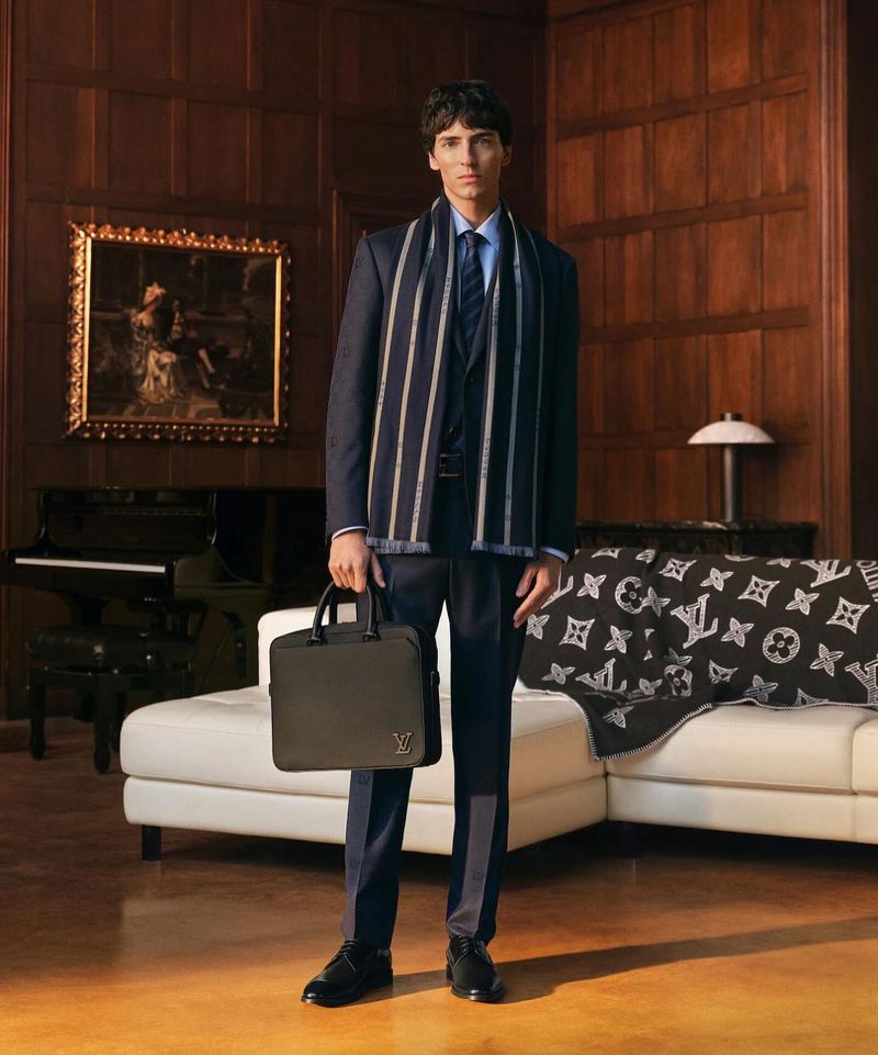 Stylish adult male in suit holds dark leather briefcase indoors. He is wearing a striped scarf, in a classically styled room with piano.