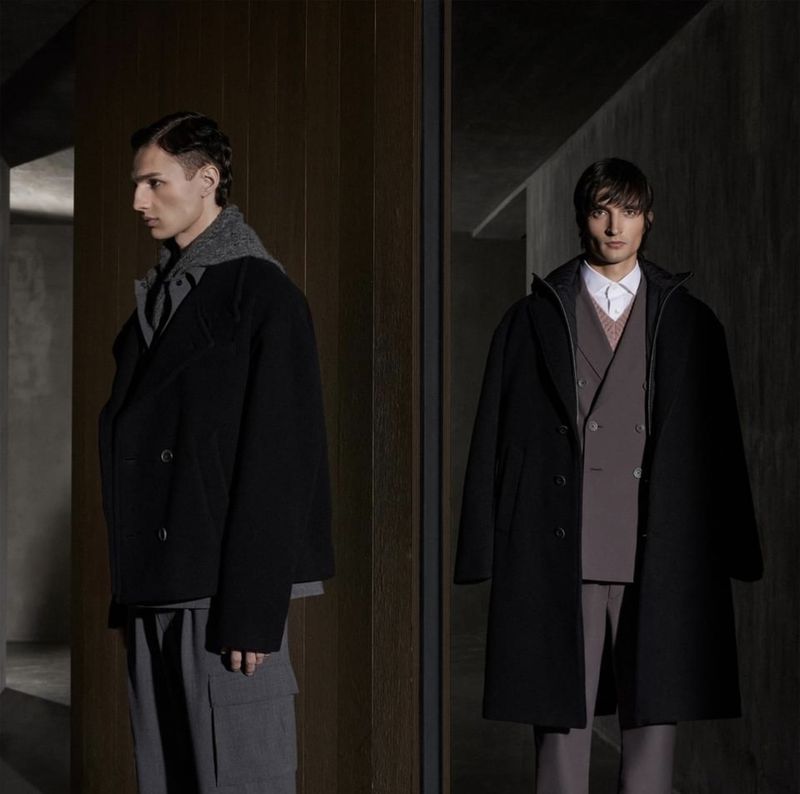 Two adult males pose in a dimly lit interior, wearing black overcoats and grey trousers, with a blurred background and studio lighting.