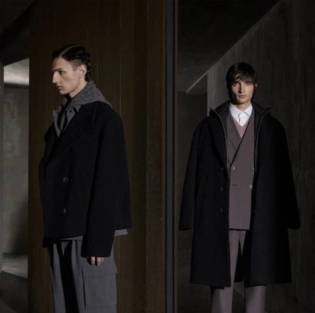 Two adult males pose in a dimly lit interior, wearing black overcoats and grey trousers, with a blurred background and studio lighting.