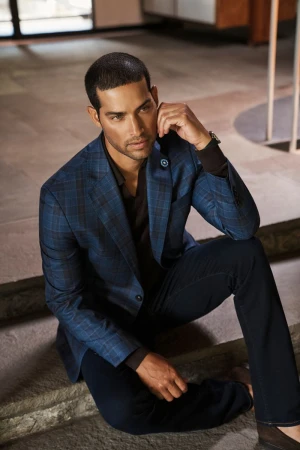 Portrait of a handsome adult male wearing a blue plaid suit, sitting on concrete steps, looking left against a blurred background.