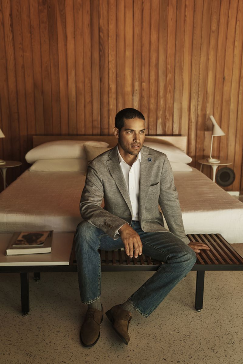 An adult male sits on a wooden bench in a bedroom interior, wearing a grey blazer, white shirt, blue jeans, and brown shoes.