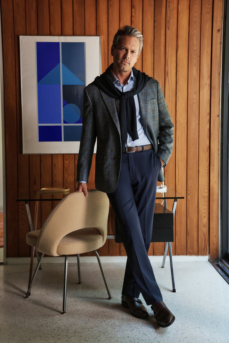 Portrait of adult man in a blazer and dress pants leaning on modern chair in front of wood paneling with geometric art in the background.