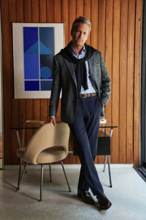 Portrait of adult man in a blazer and dress pants leaning on modern chair in front of wood paneling with geometric art in the background.