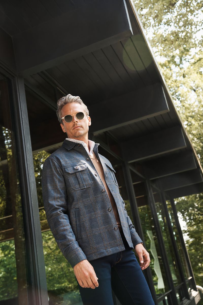A stylish adult male poses in a dark blue plaid jacket and sunglasses against a modern building and lush green trees.