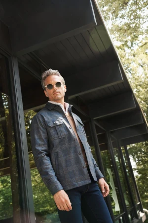 A stylish adult male poses in a dark blue plaid jacket and sunglasses against a modern building and lush green trees.