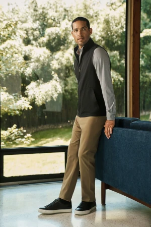 A man in a black vest and khaki pants stands near a window with a view of trees, leaning against a teal couch in a modern living room.
