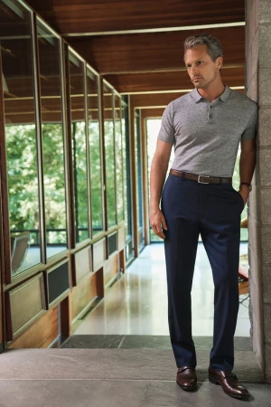 Man in polo and trousers leaning against a wall in a modern home with large windows and trees visible outside.