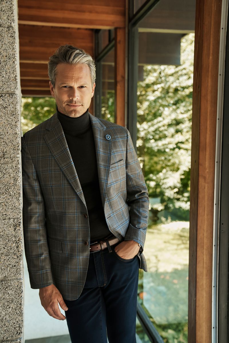 Mature man in a blazer and turtleneck stands indoors by a window; trees visible outside, confident pose, stylish, well-lit.
