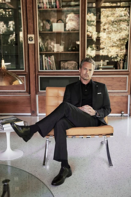 A man in a black suit poses indoors while seated on a tan chair in front of a wood paneled wall with book shelves.