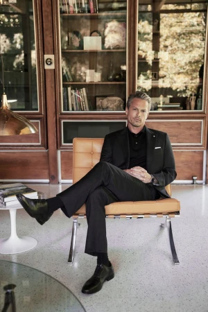A man in a black suit poses indoors while seated on a tan chair in front of a wood paneled wall with book shelves.