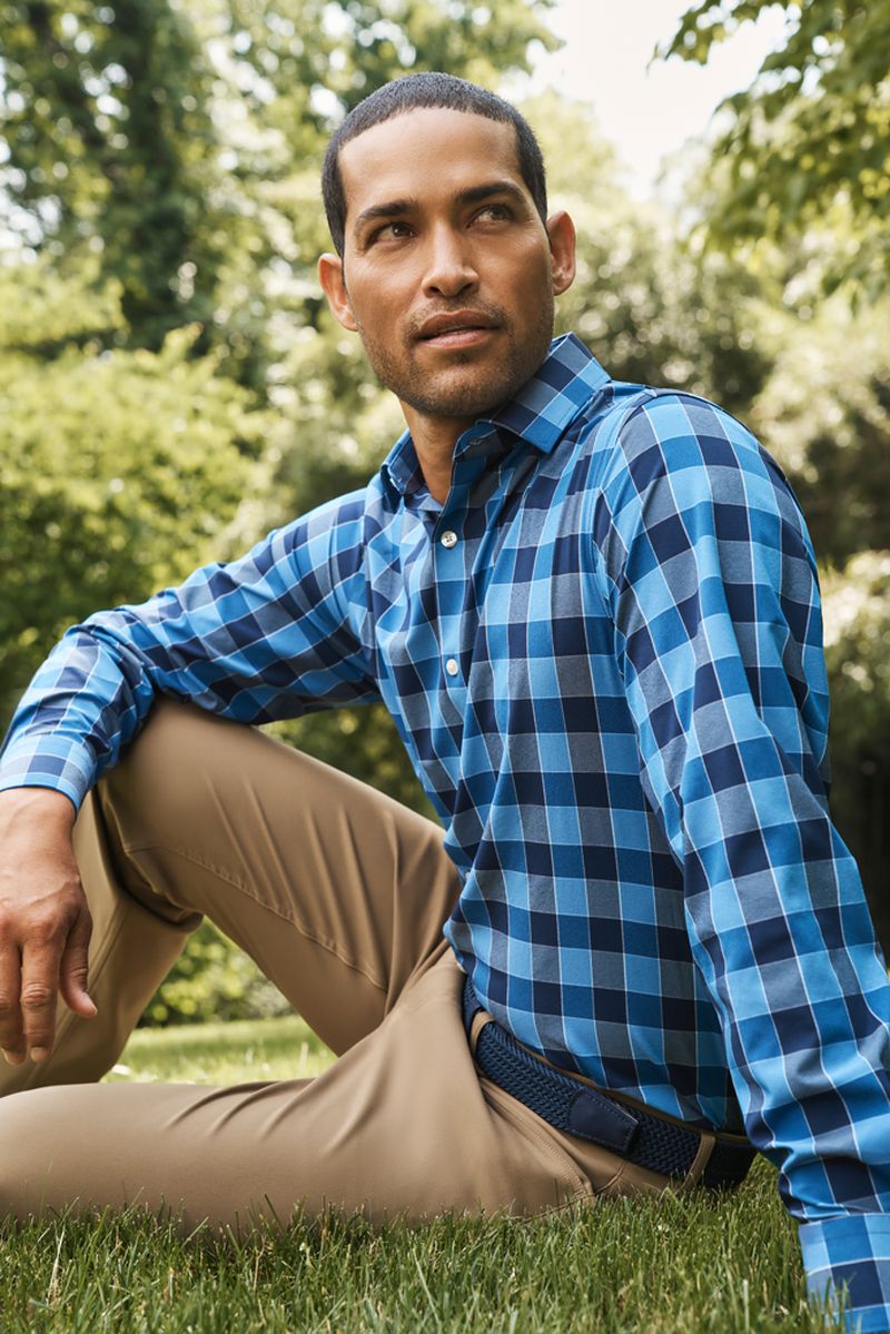 A man with short dark hair sits on green grass in a park, wearing a long-sleeved blue plaid shirt and khaki pants on a sunny day.